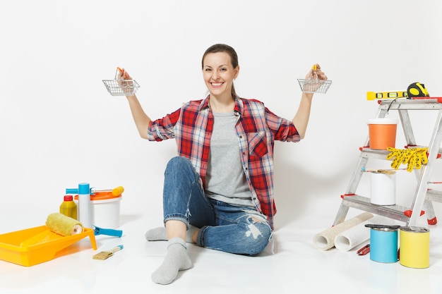 Vrouw houdt metalen boodschappenmanden om te winkelen, zit op de vloer met instrumenten voor renovatie appartement geïsoleerd op een witte achtergrond. Behang, accessoires, gereedschap om te schilderen. Reparatie huis concept.