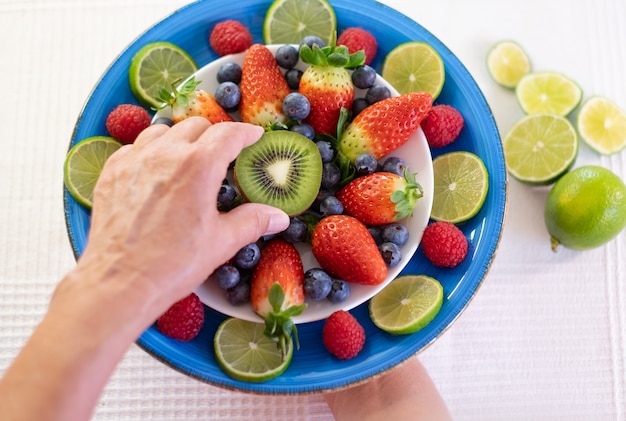 Vrouw houdt op haar hand een blauw bord vol aardbeien en bosbessen met een halve kiwi. Concept van gezonde voeding en dieet. witte achtergrond