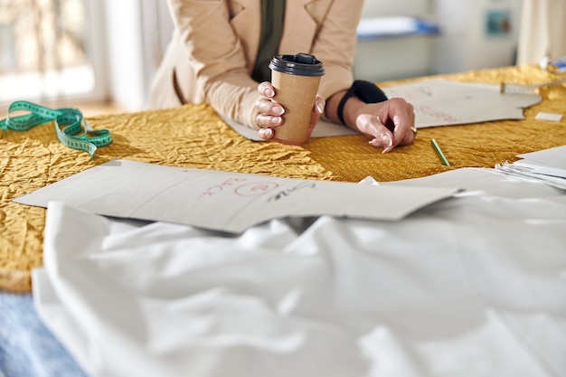 Vrouw houdt papieren beker leunend op snijtafel met stof en gereedschap in werkplaats