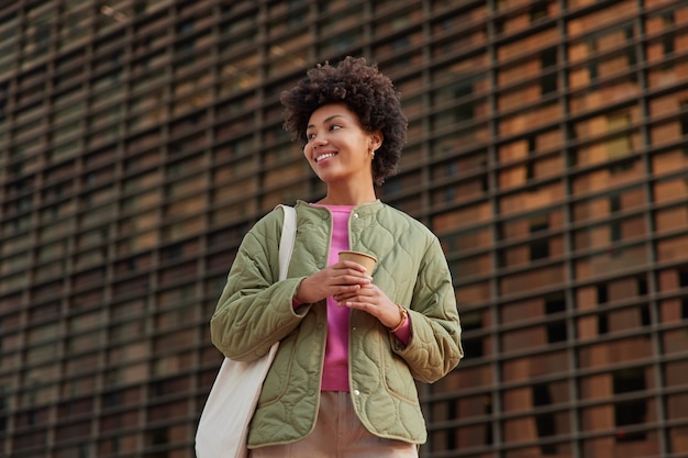 vrouw houdt papieren kopje koffie drinkt aromatische drank draagt jas draagt tas kijkt weg glimlacht vrolijk poseert tegen muur geniet van vrije tijd