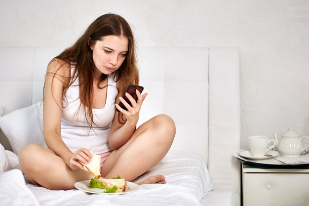 Vrouw houdt smartphone vast en eet broodjes in bed