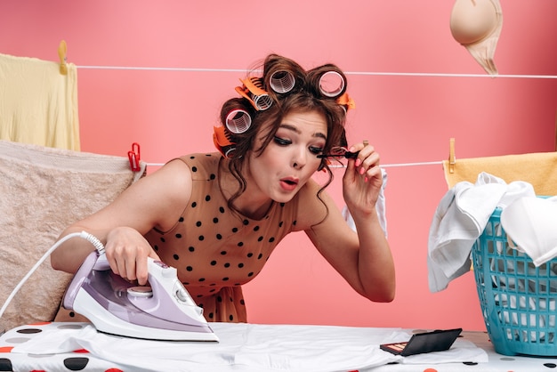 Vrouw, huisvrouw die kleren op een scherpe raad strijken en tegelijkertijd make-up doet.