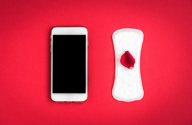 Vrouw hygiënebescherming of maandverband met rozenblaadjes en mobiele telefoon op rode achtergrond. Tracking van de menstruatiecyclus.