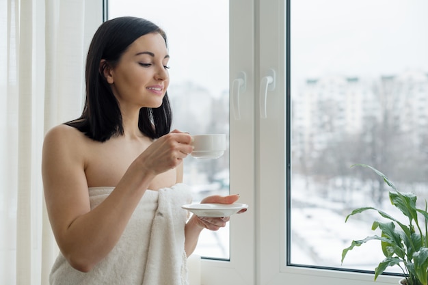 Vrouw in badhanddoek met kop van verse koffie dichtbij het venster