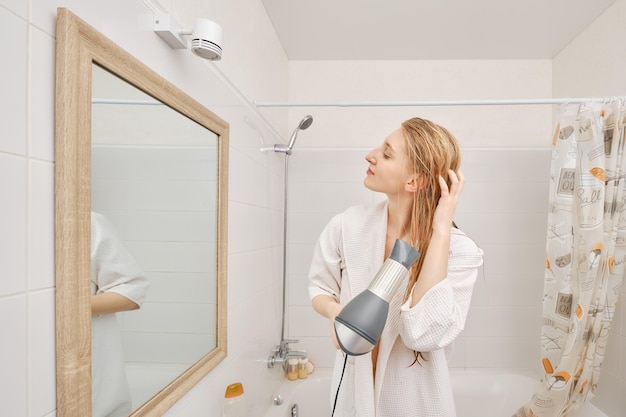 Vrouw in badjas die haar haar droogt met droger die in profiel voor de spiegel in de badkamer staat