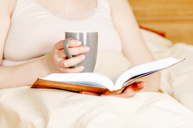 Vrouw in bed met theekop en een boek. Volwassen vrouwenlezing in het bed.