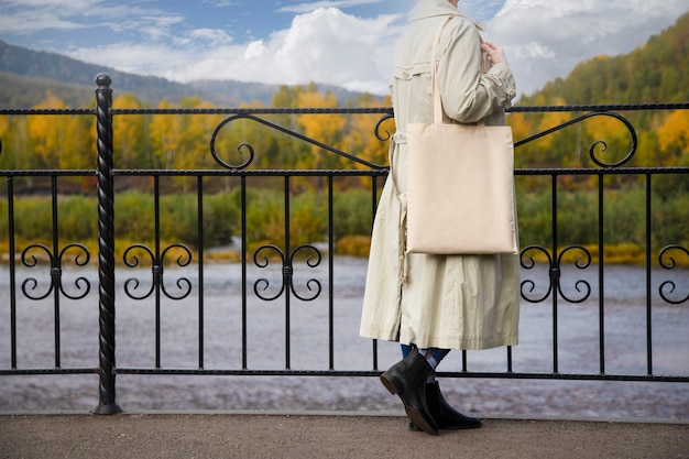 Vrouw in beige loopgraaf met lege herbruikbare boodschappentas in de buurt van brugleuningmodel