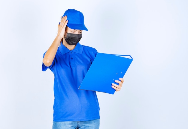 Vrouw in blauw uniform en zwart gezichtsmasker met een blauwe map en ziet er verward en attent uit.