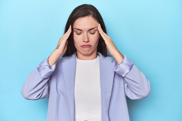 Vrouw in blauwe blazer op blauwe achtergrond die tempels aanraakt en hoofdpijn heeft