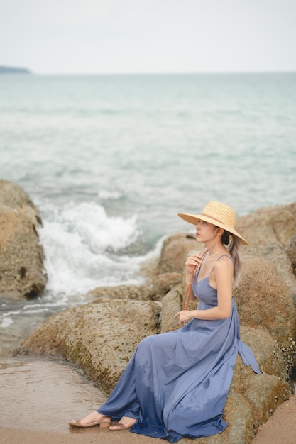 Vrouw in blauwe jurk en strohoed, zittend op een rots aan de oceaan.
