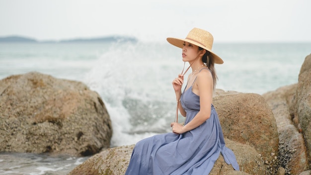 Vrouw in blauwe jurk en strohoed zittend op een rots bij de oceaan