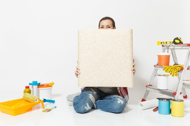 Vrouw in casual kleding zittend op de vloer met ongevouwen rol behang instrumenten voor renovatie appartement geïsoleerd op een witte achtergrond. Accessoires voor lijmgereedschap. Repareer thuis concept Kopieer ruimte