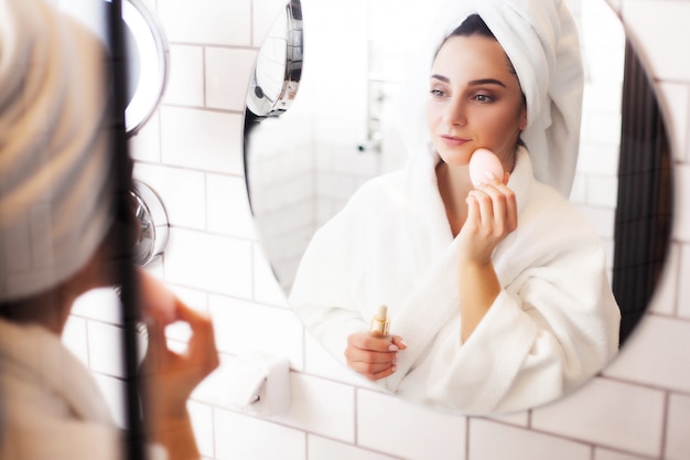 Vrouw in de badkamer met een handdoek op haar hoofd past make-up op haar gezicht