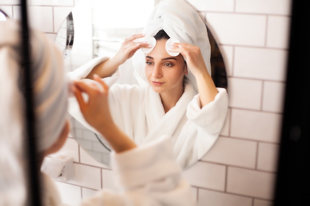 Vrouw in de badkamer met een handdoek op haar hoofd past make-up op haar gezicht