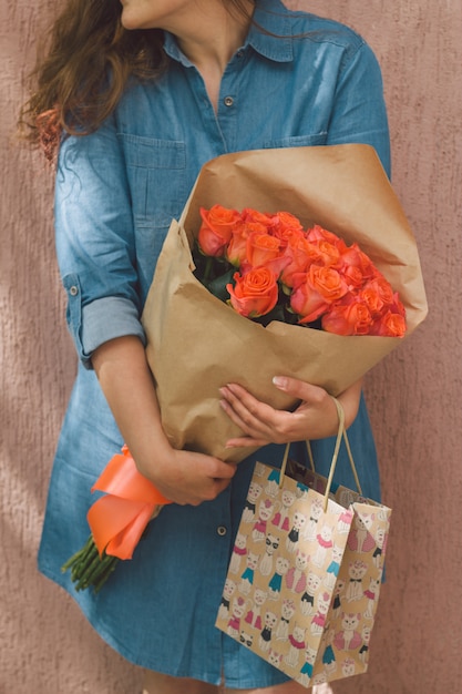 vrouw in denim jurk met boeket rozen en cadeau papieren zak