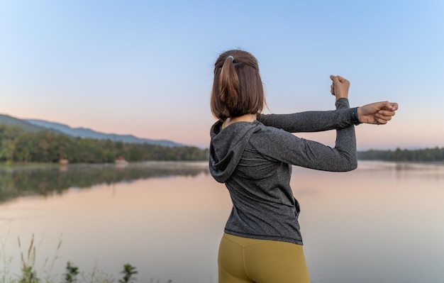 Vrouw in donkere jas die haar armen strekt tijdens haar pauze van haar ochtendoefening bij een plaatselijk meer