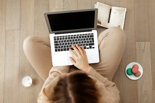 Vrouw in een beige gebreide trui werkt op de computer zittend op de vloer