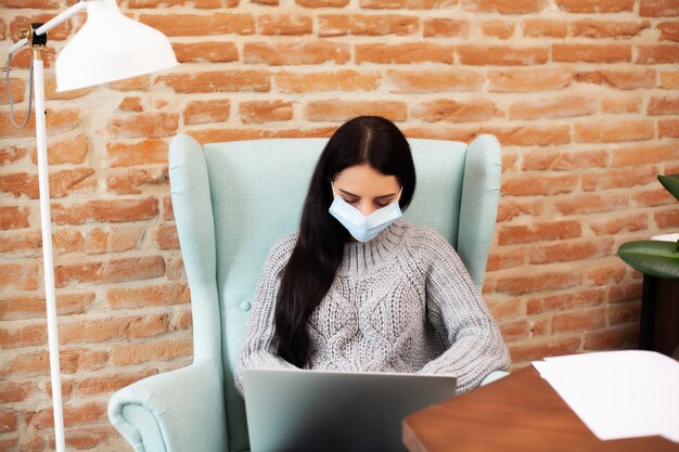 Vrouw in een beschermend masker werkt thuis op een laptop tijdens een wereldwijde epidemie van een virus