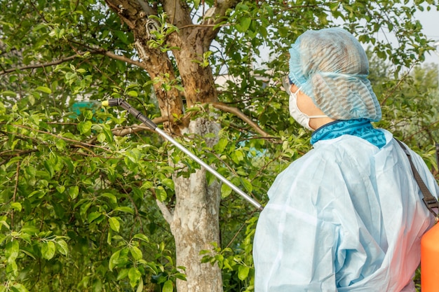 Vrouw in een beschermend pak en masker spuit appelboom tegen schimmelziekte of ongedierte met drukspuit en chemicaliën in de lenteboomgaard.