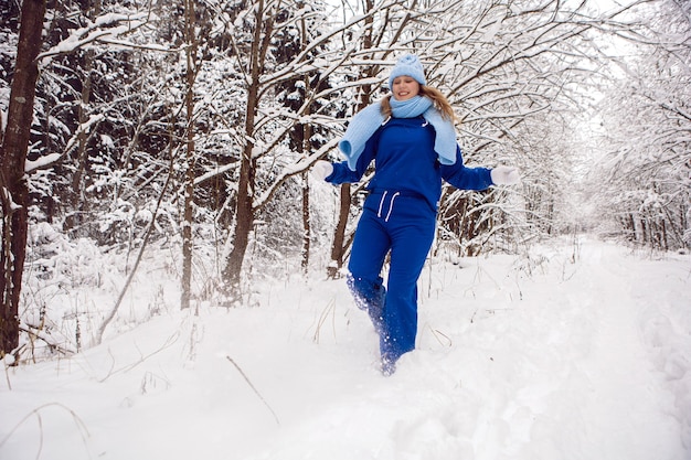 Vrouw in een blauw trainingspak witte wanten en sjaal staat in de winter in een besneeuwd bos