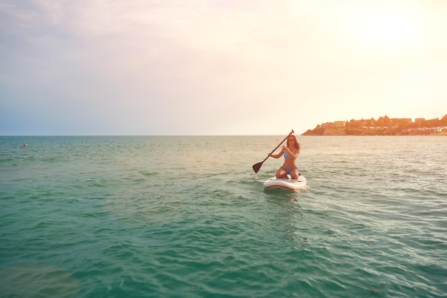 Vrouw in een blauwe bikini zit op een sup board met een peddel in de zee tegen de zonsondergang