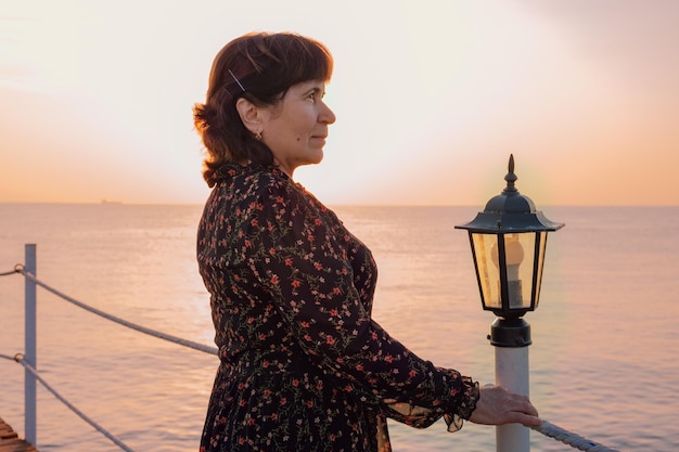 Vrouw in een donkere zomerjurk staat op een pier bij de zee tegen de achtergrond van de opkomende zon