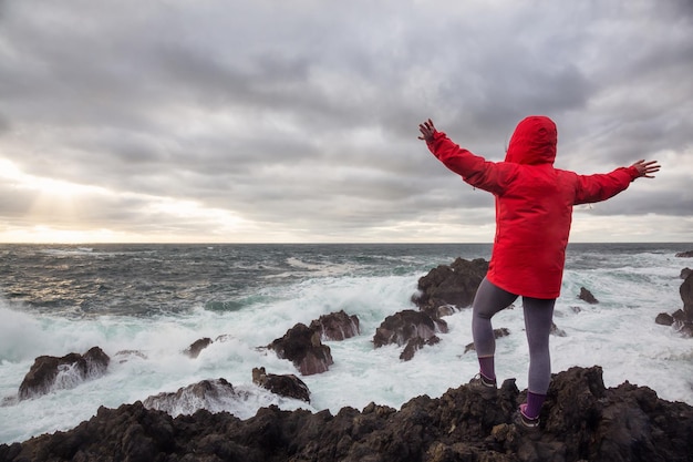 Vrouw in een felrood jasje kijkt naar golven die neerstorten op een rotsachtige kust van de Stille Oceaan