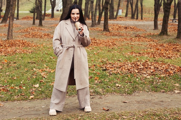 Vrouw in een grijze jas wandelen in het herfstpark met een kop warme drank