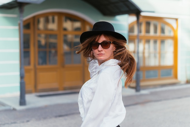 Vrouw in een hoed en zonnebril die in de zomer met haar haar in de stad zwaait