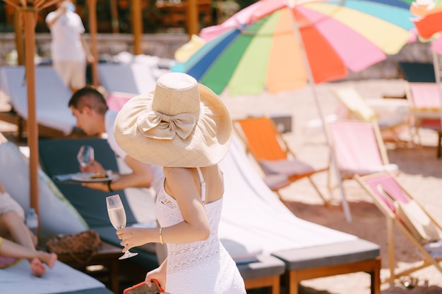 Vrouw in een hoed met een glas in haar hand loopt langs het strand achteraanzicht