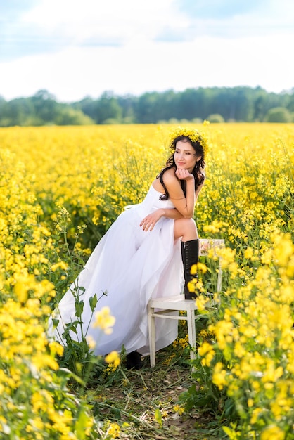 Vrouw in een lange witte jurk en zwarte laarzen zit in een koolzaadveld het meisje geniet van de natuur en vrijheid