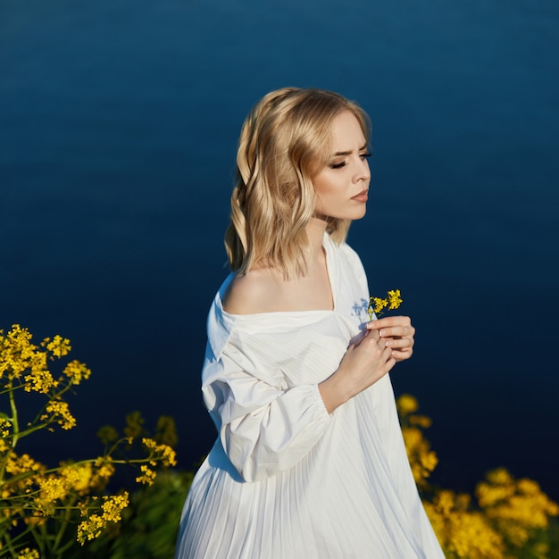 vrouw in een lange witte jurk met een bloem in haar hand staat in de buurt van het meer. Blondevrouw in de zon in een lichte kleding. vrouw rusten en dromen, perfecte zomer make-up