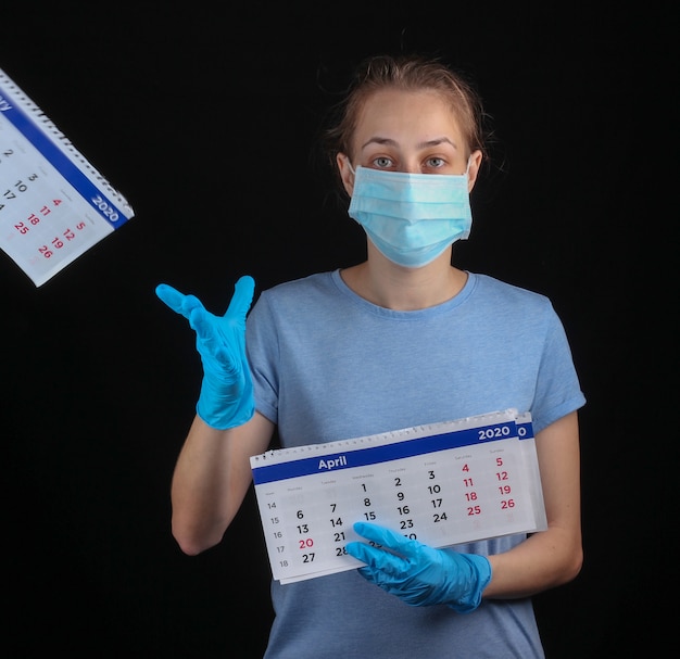 Vrouw in een medisch beschermend masker, handschoenen houden maandkalender op een zwarte muur. Quarantaine, pandemische covid-19