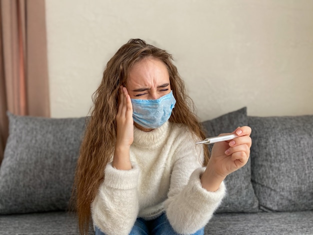 Vrouw in een medisch masker zit thuis met een thermometer in haar handen en hoofdpijn