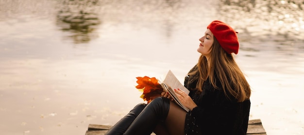 Vrouw in een rode baret leesboek op houten ponton Herfstseizoen Vrouw met trendy kleding ontspannen in de buurt van water leesboek en koffie drinken Herfststemming Herfstconcept Herfstdag