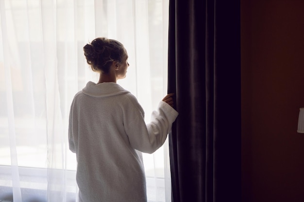 Vrouw in een wit gewaad staat na een douche op een bed bij het raam in een hotelappartement