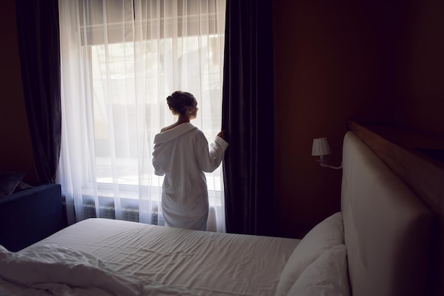 Vrouw in een wit gewaad staat na een douche op een bed bij het raam in een hotelappartement