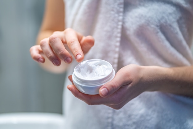 Vrouw in een witte badhanddoek die voor haar huid zorgt en hydraterende en voedende lichaamscrème na bad thuis gebruikt. Huidverzorgingsproduct