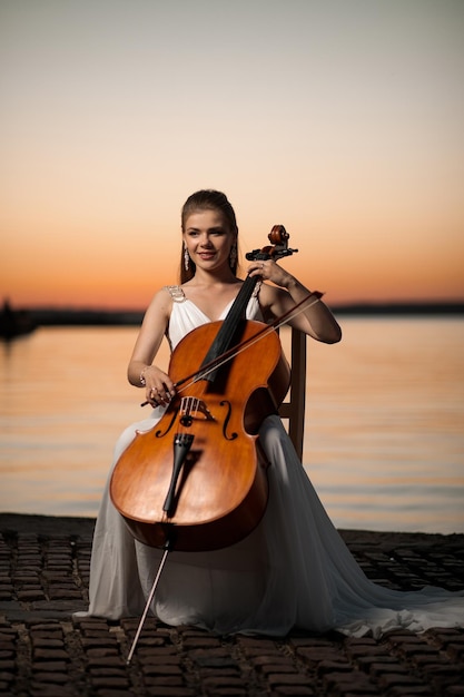 Vrouw in een witte jurk die cello speelt aan de oever van het meer na zonsondergang