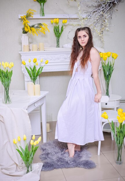 vrouw in een witte jurk zit op een stoel en glimlacht gelukkig In de voorkant is er een vaas met gele tulpen