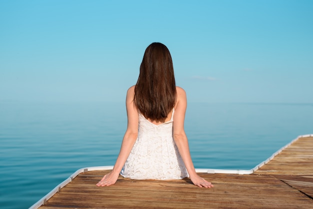 vrouw in een witte jurk zittend op de pier en op zoek in de verte