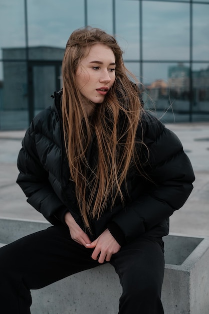 Vrouw in een zwart kort donsjack en joggingbroek zittend op een betonnen stoeprand Streetstyle dame die een capuchon op doet