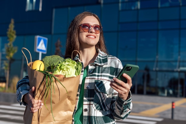 Vrouw in geel jasje met smartphone en boodschappentas met boodschappen in handen