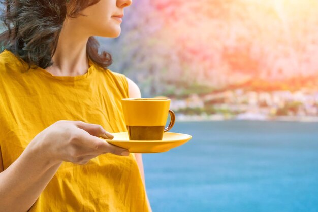 Vrouw in gele kleren houdt een gele en bruine beker met koffie tegen de zee
