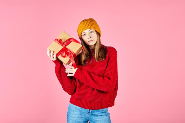 Vrouw in gezellige rode trui geschenkdozen in handen houden en cadeau kiezen op roze achtergrond Besluiteloos jong hipster meisje met gebreide muts die een geschenkdoos kiest