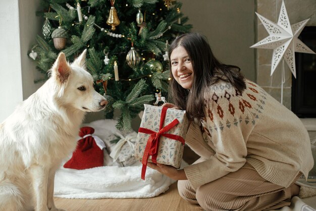 Vrouw in gezellige trui kerstcadeau openen met schattige witte hond op achtergrond van stijlvolle kerstboom en open haard Wintervakantie Eigenaar met huisdier vieren in feestelijke kamer Vrolijk kerstfeest