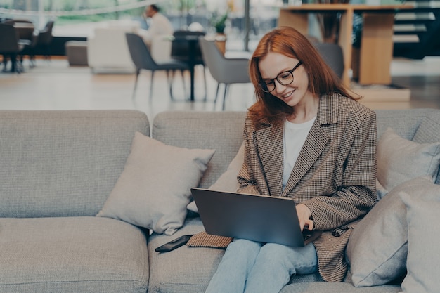 Vrouw in glazen zittend op een gezellige bank in de kantoorlounge tijdens het chatten op een computerlaptop