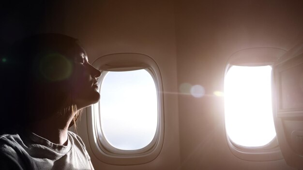 Vrouw in grijs t-shirt half gezicht silhouet tegen heldere ramen van het passagiersvliegtuig rustend in de passagierscabine van dichtbij bekeken
