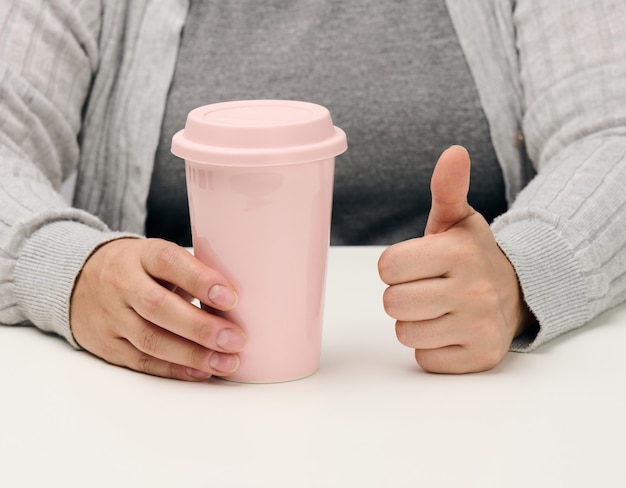 Vrouw in grijze kleding houdt een roze keramische beker vast, vrouw zit aan een witte tafel, begin van de dag, ochtend