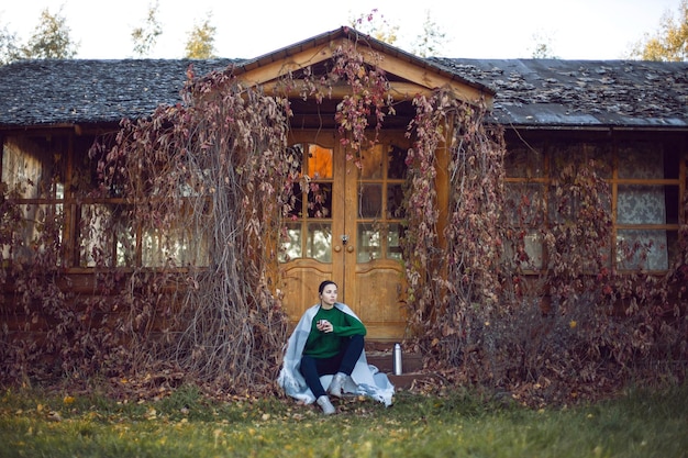 Foto vrouw in groene trui en plaid zit op veranda met klimop in de herfst en drinkt thee uit een thermos